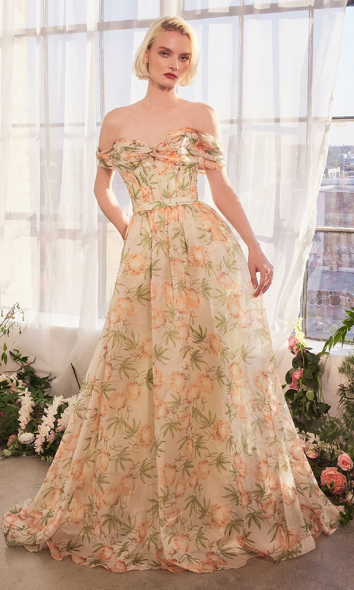 A woman stands indoors wearing the Andrea & Leo A1466 Long Floral-Print Prom Dress, featuring an off-the-shoulder corset-style bodice and flowing skirt. Flowers and greenery are arranged on the floor around her.